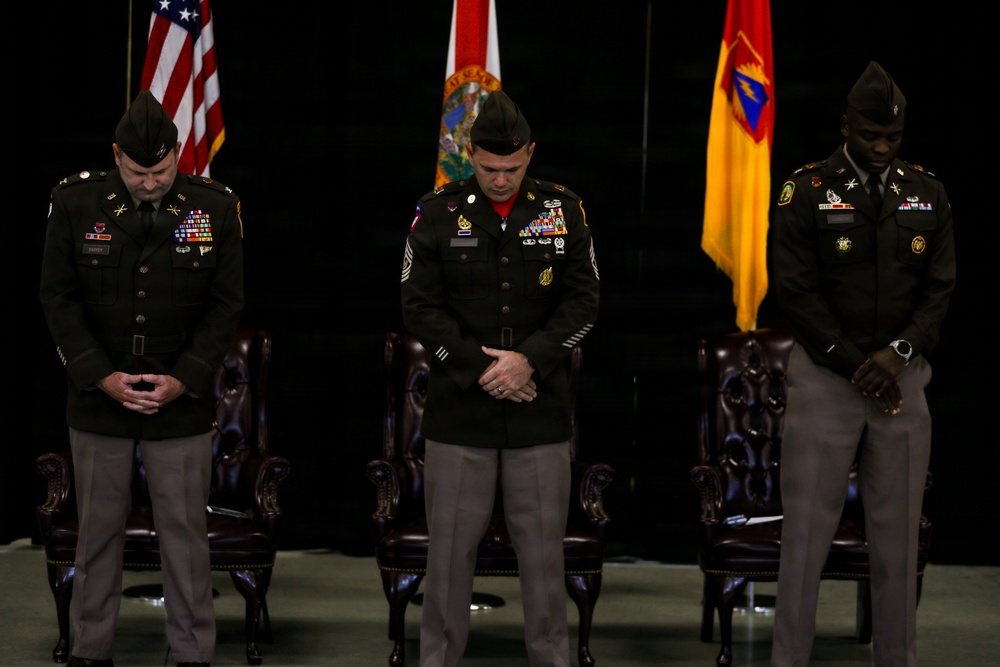 Three U.S. Army soldiers in dress green uniforms stand with heads bowed in what appears to be a formal military ceremony, likely a prayer or moment of reflection. They are positioned in front of multiple flags including the U.S. flag and what appears to be the Florida state flag and a unit flag. The setting appears to be an indoor ceremonial venue with ornate leather chairs visible behind them.