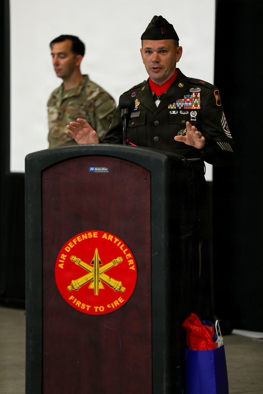 A senior U.S. Army non-commissioned officer in dress greens uniform is speaking at a podium bearing the Air Defense Artillery 'First to Fire' insignia. He is wearing a Medal of Honor on a red ribbon around his neck, along with numerous service ribbons, badges, and sergeant major rank insignia. A second soldier in Army Combat Uniform (ACU/OCP) stands in the background.