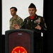 A senior U.S. Army non-commissioned officer in dress greens uniform is speaking at a podium bearing the Air Defense Artillery 'First to Fire' insignia. He is wearing a Medal of Honor on a red ribbon around his neck, along with numerous service ribbons, badges, and sergeant major rank insignia. A second soldier in Army Combat Uniform (ACU/OCP) stands in the background.