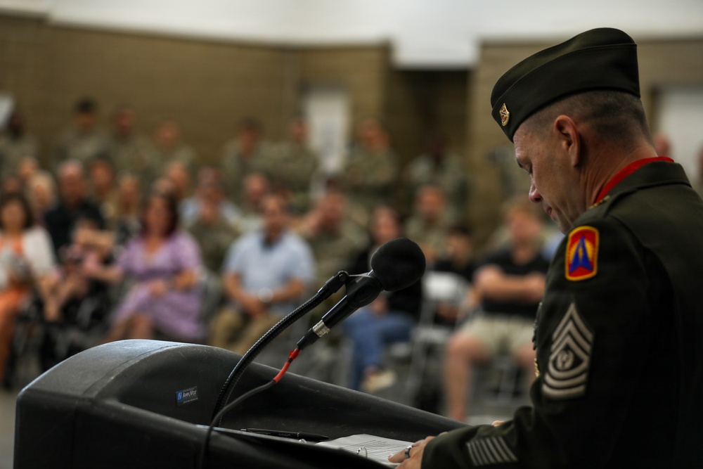 A senior non-commissioned officer in U.S. Army dress green uniform stands at a podium addressing a seated audience during what appears to be a formal military ceremony. The speaker is viewed from behind and to the side, with a microphone visible at the podium. The background shows a large crowd of both military personnel and civilians seated in rows.