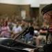 A senior non-commissioned officer in U.S. Army dress green uniform stands at a podium addressing a seated audience during what appears to be a formal military ceremony. The speaker is viewed from behind and to the side, with a microphone visible at the podium. The background shows a large crowd of both military personnel and civilians seated in rows.