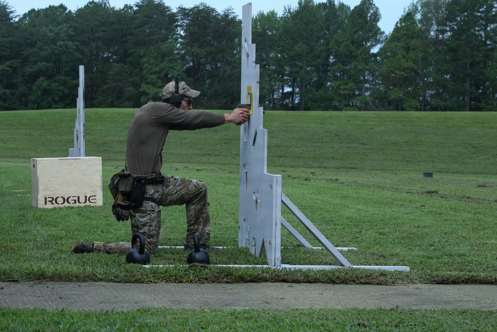 A person in military/tactical gear is kneeling on a grass field and aiming a pistol through a gap in a white barricade structure at a shooting range. The shooter is wearing camouflage pants, a brown long-sleeve shirt, a cap, and hearing protection. Several range props including kettlebells and a Rogue-branded wooden box are visible nearby.