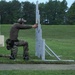 A person in military/tactical gear is kneeling on a grass field and aiming a pistol through a gap in a white barricade structure at a shooting range. The shooter is wearing camouflage pants, a brown long-sleeve shirt, a cap, and hearing protection. Several range props including kettlebells and a Rogue-branded wooden box are visible nearby.