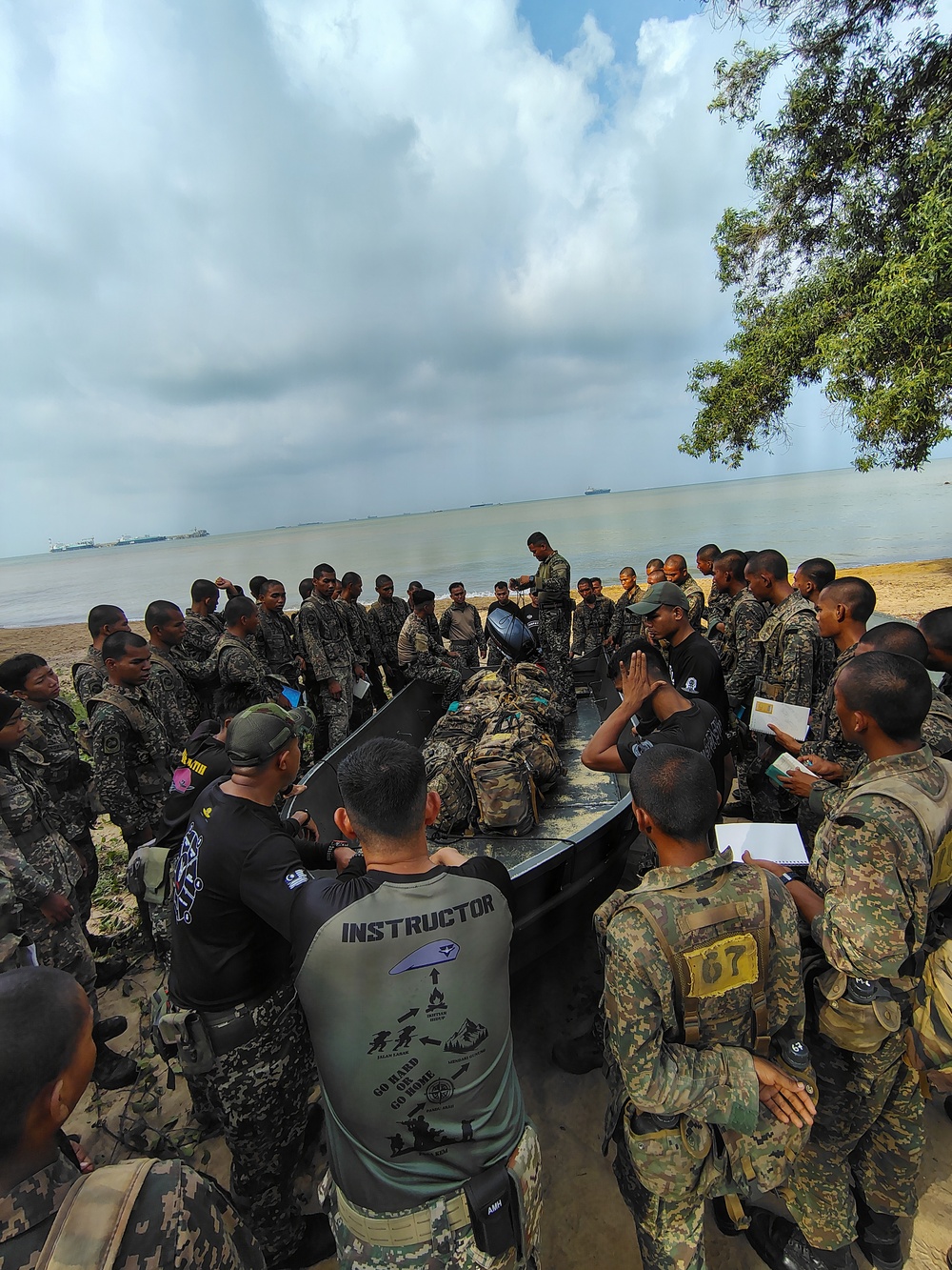 A military training session is taking place on a beach, with an instructor demonstrating techniques to a large group of soldiers in camouflage uniforms. A person in full gear is lying prone on what appears to be a dark-colored inflatable boat or rubber craft, likely serving as a demonstration subject. The setting appears to be a coastal training area with ships visible on the horizon.