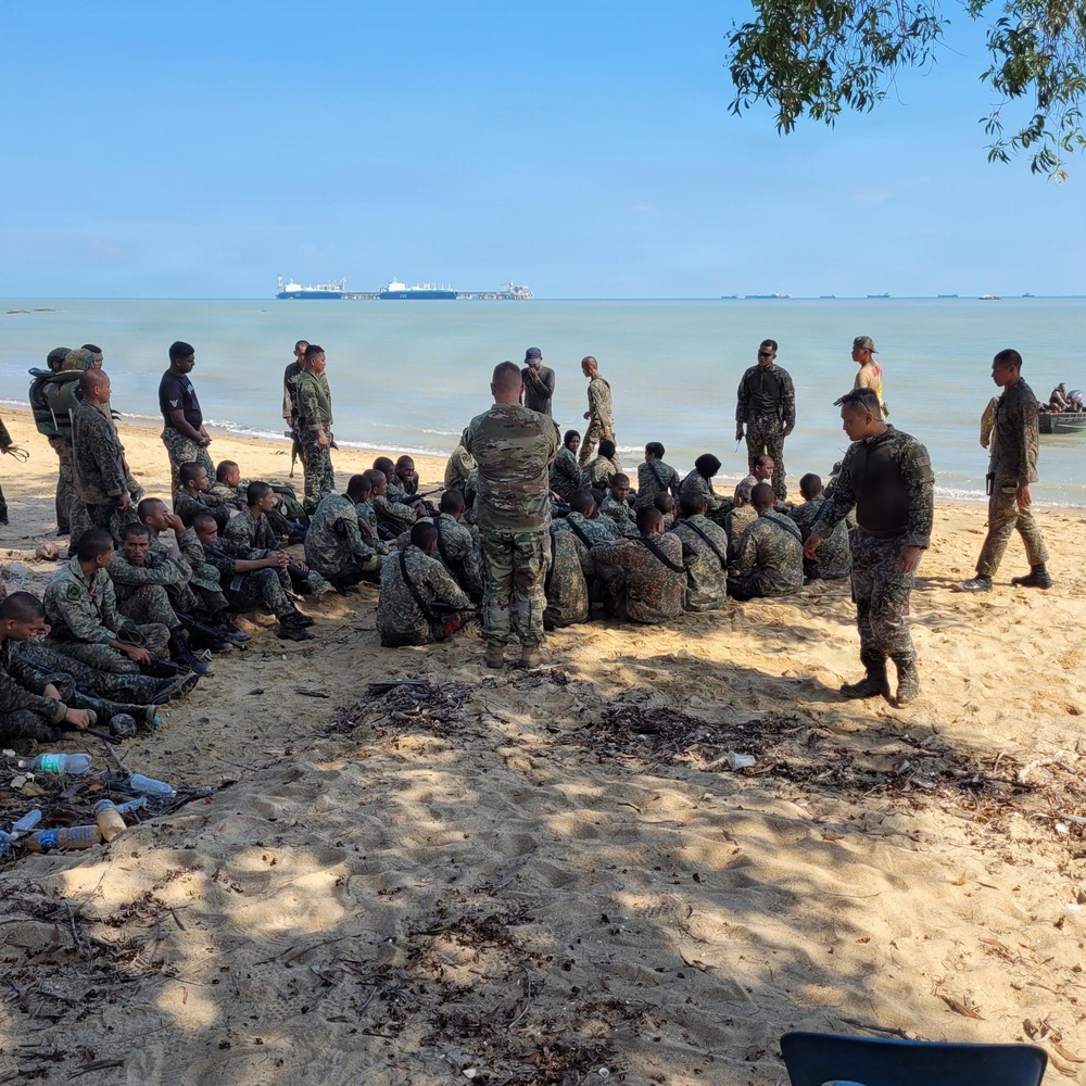 A group of military or paramilitary personnel are gathered on a sandy beach, with a mix of individuals seated and standing. An instructor or leader figure in multicam/OCP camouflage appears to be conducting a briefing or training session for a larger group in matching camouflage uniforms. Large cargo or tanker vessels are visible on the horizon in the background.