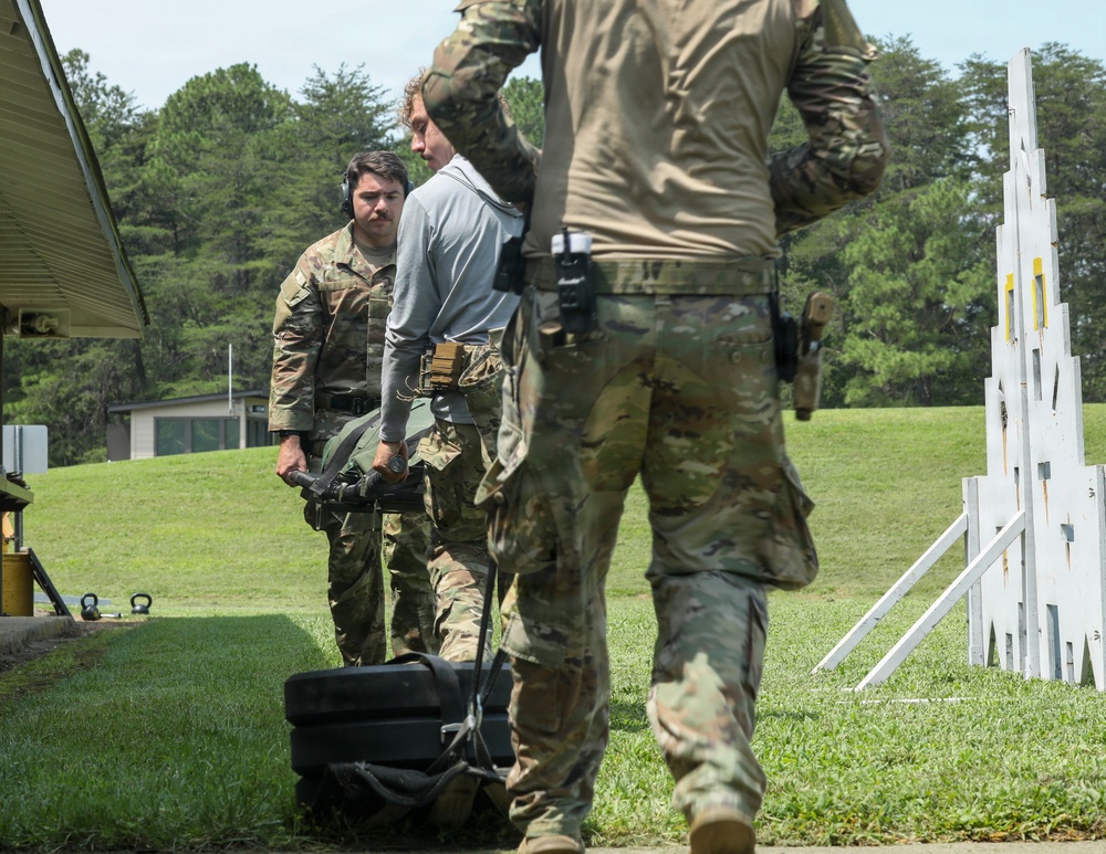 The image shows military personnel at what appears to be an outdoor shooting range or training facility. Several individuals in camouflage uniforms are engaged in a training exercise, with one person handling what appears to be a weapon or equipment near a black case. White target stands with yellow markings are visible in the background on a grassy field.