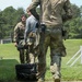 The image shows military personnel at what appears to be an outdoor shooting range or training facility. Several individuals in camouflage uniforms are engaged in a training exercise, with one person handling what appears to be a weapon or equipment near a black case. White target stands with yellow markings are visible in the background on a grassy field.