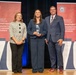 Three individuals are posing for a formal photograph on a stage at what appears to be an awards ceremony for the Department of War Manufacturing Technology Program. The central figure is holding a crystal/glass award trophy, suggesting they are the recipient of a recognition. The event appears to be celebrating the program's 70th anniversary, as indicated by the large '70' numerals visible on either side of the backdrop.