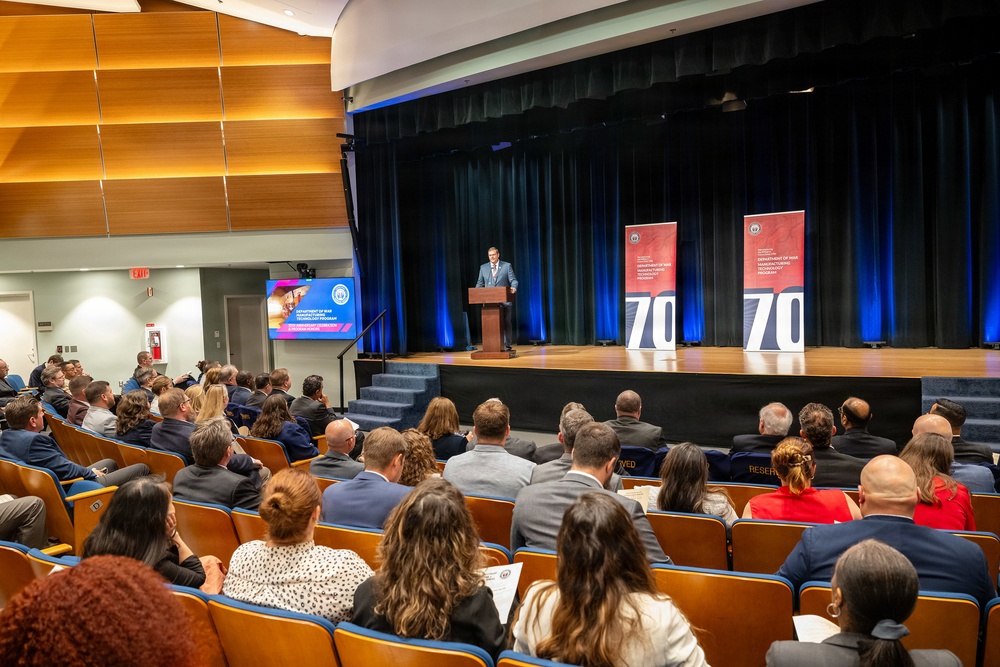 A formal event is taking place in a modern auditorium where a speaker addresses a seated audience from a podium on a raised stage. Two red banner stands displaying the number '70' flank the stage, along with a digital display screen showing what appears to be 'Department of War Manufacturing Technology Program' branding. The audience, dressed in business attire, fills the theater-style seating.