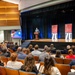 A formal event is taking place in a modern auditorium where a speaker addresses a seated audience from a podium on a raised stage. Two red banner stands displaying the number '70' flank the stage, along with a digital display screen showing what appears to be 'Department of War Manufacturing Technology Program' branding. The audience, dressed in business attire, fills the theater-style seating.