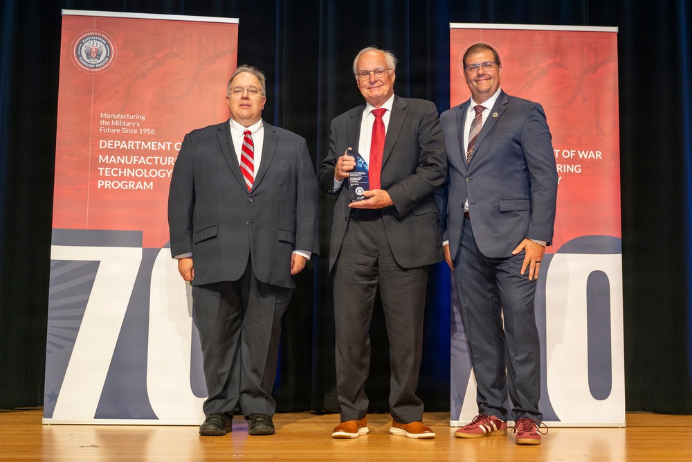Three men in business attire are posing on a stage during an award ceremony. The center individual is holding a crystal or glass award trophy. The backdrop features banners for the Department of Defense Manufacturing Technology Program, celebrating its history since 1956.