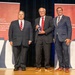 Three men in business attire are posing on a stage during an award ceremony. The center individual is holding a crystal or glass award trophy. The backdrop features banners for the Department of Defense Manufacturing Technology Program, celebrating its history since 1956.
