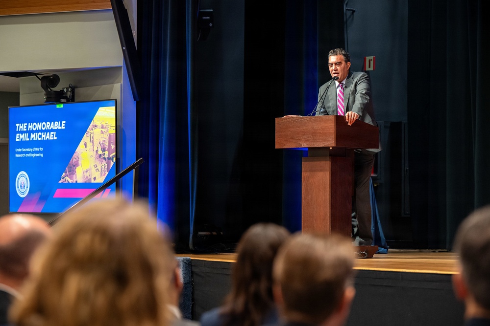 A man identified as 'The Honorable Emil Michael, Under Secretary of War for Research and Engineering' is delivering a speech at a podium on a raised stage. He is wearing a gray suit with a pink/striped tie and speaking into a microphone. An audience is visible in the foreground, and a presentation slide displaying his name and title is shown on a screen to his left.