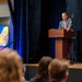 A man identified as 'The Honorable Emil Michael, Under Secretary of War for Research and Engineering' is delivering a speech at a podium on a raised stage. He is wearing a gray suit with a pink/striped tie and speaking into a microphone. An audience is visible in the foreground, and a presentation slide displaying his name and title is shown on a screen to his left.