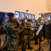 A military brass band is rehearsing or performing indoors, with soldiers in camouflage uniforms and blue berets playing various brass instruments. The group includes both male and female musicians arranged in formation with music stands. The setting appears to be a large indoor hall or gymnasium with white walls and blue-framed windows.