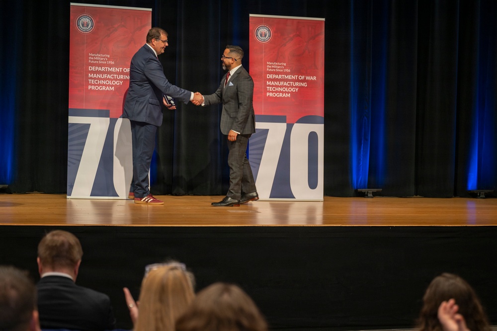 Two men in business suits are shaking hands on a stage during what appears to be an awards ceremony. The event is associated with the Department of War Manufacturing Technology Program, with banners displaying '70' suggesting a 70th anniversary celebration. An audience is visible in the foreground watching the presentation.