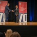 Two men in business suits are shaking hands on a stage during what appears to be an awards ceremony. The event is associated with the Department of War Manufacturing Technology Program, with banners displaying '70' suggesting a 70th anniversary celebration. An audience is visible in the foreground watching the presentation.