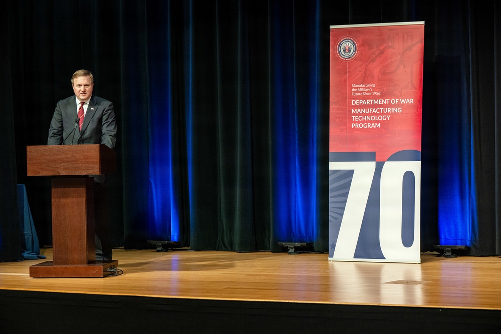 A man in a dark suit with a red tie is speaking at a wooden podium on a stage. Behind him is a dark curtain backdrop with blue lighting, and to his right stands a pull-up banner for the 'Department of War Manufacturing Technology Program' celebrating its 70th anniversary, with the tagline 'Manufacturing the Military's Future Since 1956.'
