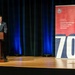 A man in a dark suit with a red tie is speaking at a wooden podium on a stage. Behind him is a dark curtain backdrop with blue lighting, and to his right stands a pull-up banner for the 'Department of War Manufacturing Technology Program' celebrating its 70th anniversary, with the tagline 'Manufacturing the Military's Future Since 1956.'