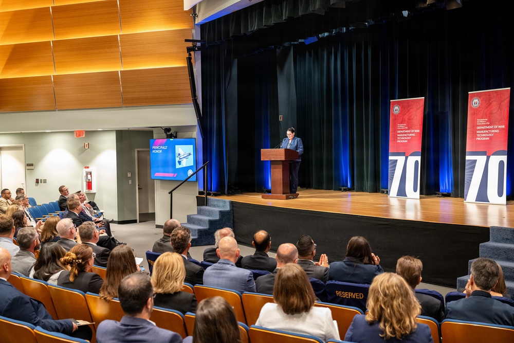 A formal ceremony or presentation is taking place in a modern auditorium, where a speaker stands at a wooden podium on a raised stage. Two red banner stands displaying '70' and text reading 'Department of War Manufacturing Technology Program' flank the stage, suggesting a 70th anniversary event. An audience of professionally dressed attendees fills the seating area, with a presentation slide reading 'Principals' Honor' visible on a side screen.