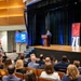 A formal ceremony or presentation is taking place in a modern auditorium, where a speaker stands at a wooden podium on a raised stage. Two red banner stands displaying '70' and text reading 'Department of War Manufacturing Technology Program' flank the stage, suggesting a 70th anniversary event. An audience of professionally dressed attendees fills the seating area, with a presentation slide reading 'Principals' Honor' visible on a side screen.