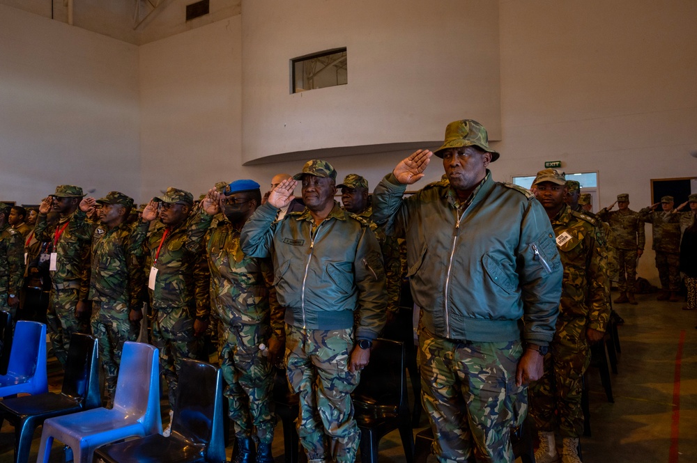 A group of military personnel in camouflage uniforms are standing at attention and saluting in what appears to be an indoor ceremonial setting. The individuals are wearing matching woodland/Portuguese-pattern camouflage uniforms, with two senior officers in the foreground also wearing olive drab flight-style jackets. The setting appears to be a formal military ceremony or official gathering.