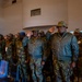 A group of military personnel in camouflage uniforms are standing at attention and saluting in what appears to be an indoor ceremonial setting. The individuals are wearing matching woodland/Portuguese-pattern camouflage uniforms, with two senior officers in the foreground also wearing olive drab flight-style jackets. The setting appears to be a formal military ceremony or official gathering.