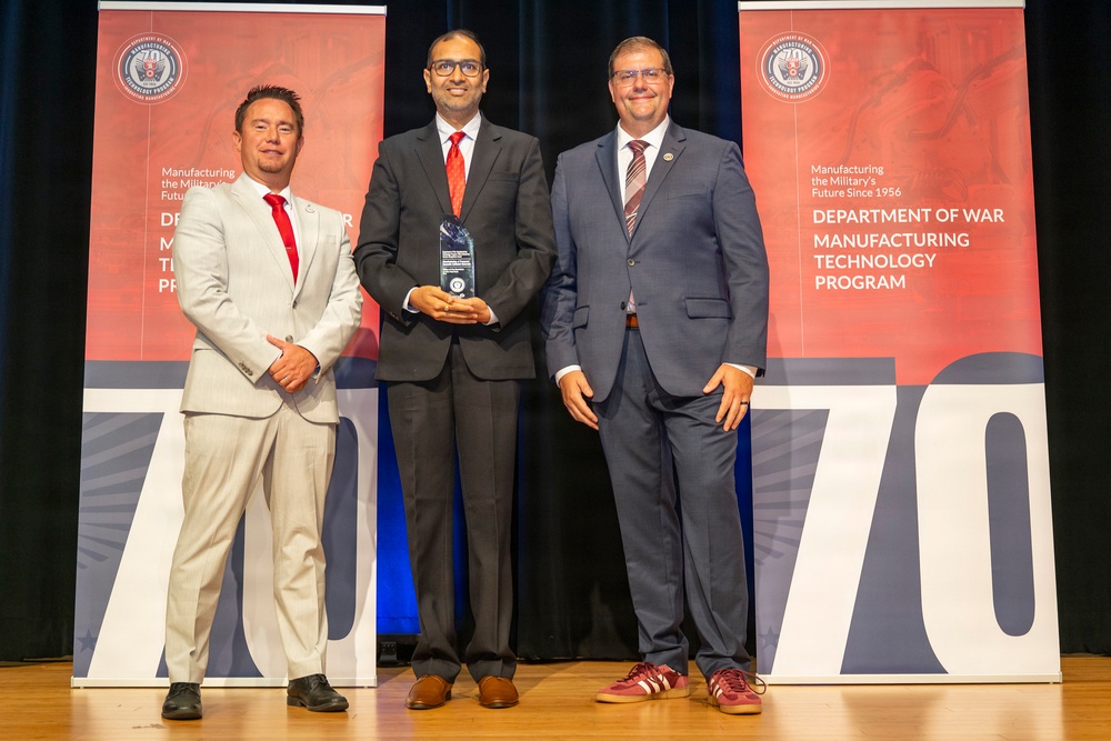 Three men in formal business attire pose on a stage at what appears to be an award ceremony for the Department of War Manufacturing Technology Program. The central figure holds a crystal/glass trophy award. The backdrop features red and white banners marking what appears to be the program's 70th anniversary.