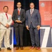Three men in formal business attire pose on a stage at what appears to be an award ceremony for the Department of War Manufacturing Technology Program. The central figure holds a crystal/glass trophy award. The backdrop features red and white banners marking what appears to be the program's 70th anniversary.