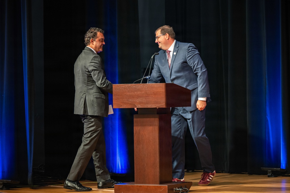 Two men in business suits are on a stage near a wooden podium with a microphone, appearing to greet or shake hands. The setting suggests a formal event, conference, or ceremony taking place in an auditorium with dark curtains and blue accent lighting. One man wears a gray suit with black dress shoes, while the other wears a navy blue suit with distinctive red/maroon athletic sneakers.