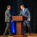 Two men in business suits are on a stage near a wooden podium with a microphone, appearing to greet or shake hands. The setting suggests a formal event, conference, or ceremony taking place in an auditorium with dark curtains and blue accent lighting. One man wears a gray suit with black dress shoes, while the other wears a navy blue suit with distinctive red/maroon athletic sneakers.