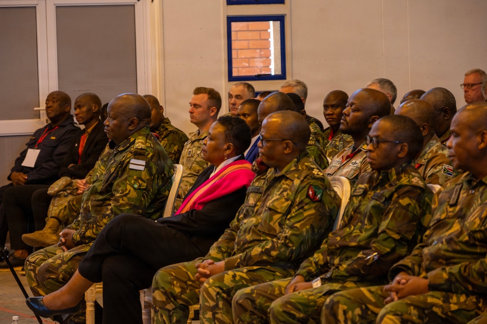 A formal indoor gathering featuring a large group of military personnel in camouflage uniforms seated in rows, alongside a small number of civilians. The attendees appear to be listening to a presentation or briefing, suggesting a multinational military meeting or conference. Multiple different uniform patterns and national insignia are visible, indicating participation from several different nations.