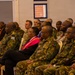 A formal indoor gathering featuring a large group of military personnel in camouflage uniforms seated in rows, alongside a small number of civilians. The attendees appear to be listening to a presentation or briefing, suggesting a multinational military meeting or conference. Multiple different uniform patterns and national insignia are visible, indicating participation from several different nations.