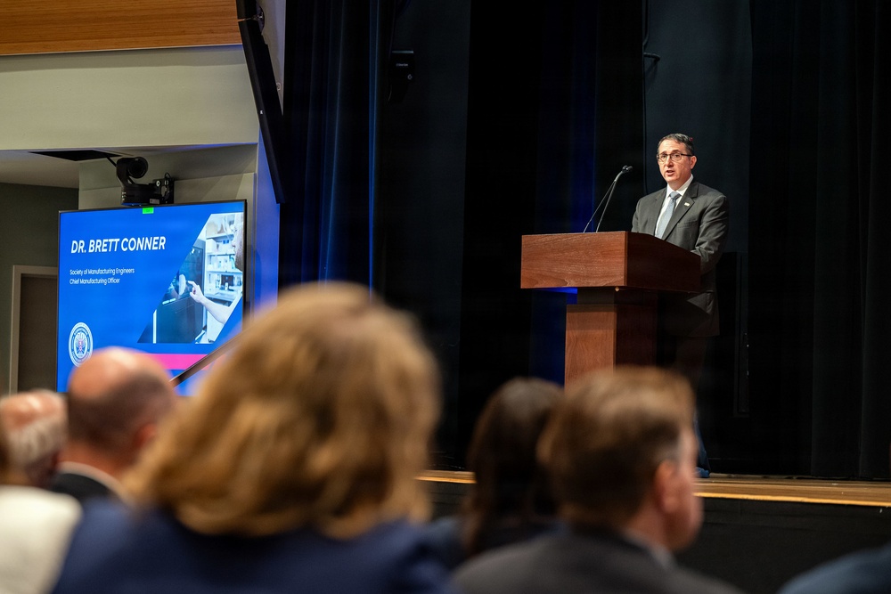 A formal presentation or conference is taking place in an auditorium-style venue. A man identified on the slide as Dr. Brett Conner, Chief Manufacturing Officer of the Society of Manufacturing Engineers, is speaking at a wooden podium on a stage. An audience is seated in the foreground, viewed from behind.