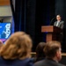 A formal presentation or conference is taking place in an auditorium-style venue. A man identified on the slide as Dr. Brett Conner, Chief Manufacturing Officer of the Society of Manufacturing Engineers, is speaking at a wooden podium on a stage. An audience is seated in the foreground, viewed from behind.