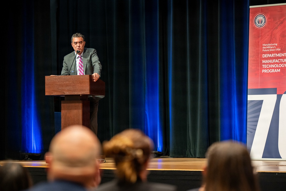 A man in a gray suit and striped tie is speaking at a wooden podium on a stage before an audience. The event appears to be associated with the Department of Defense Manufacturing Technology Program, as indicated by the banner visible on the right side of the stage. The setting suggests a formal conference or anniversary celebration, with blue stage lighting and dark curtains in the background.
