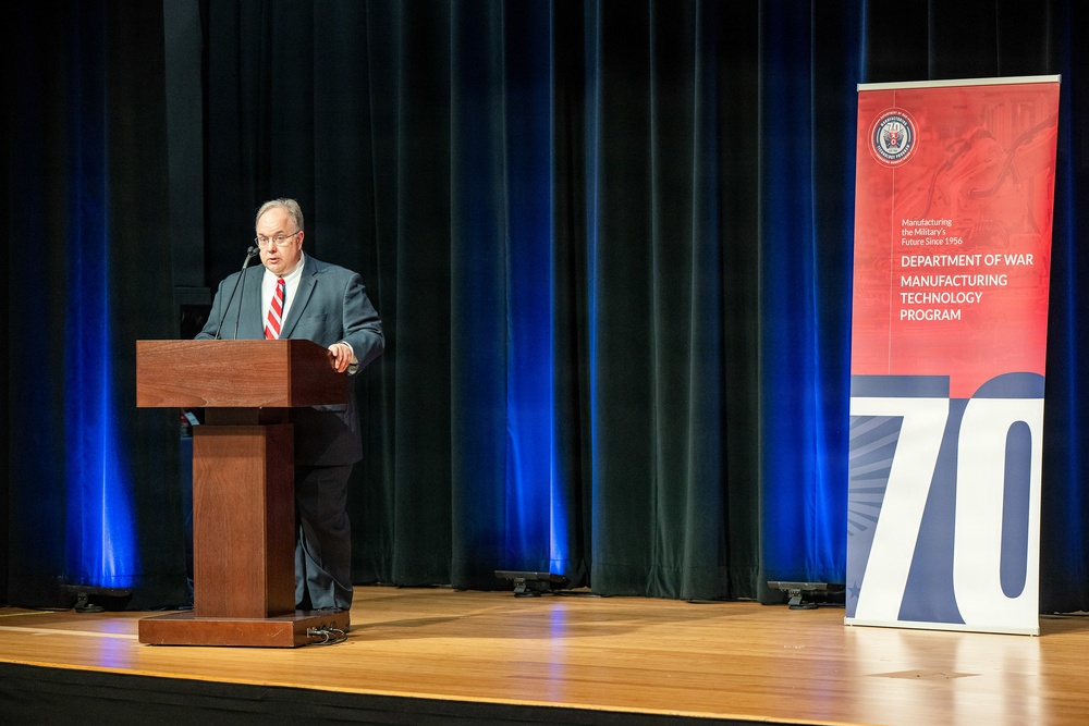 A man in a dark suit and red striped tie is speaking at a wooden podium on a stage. Behind him is a dark teal curtain with blue stage lighting, and to his right stands a banner for the 'Department of War Manufacturing Technology Program' marking its 70th anniversary. The event appears to be a formal ceremony or conference.