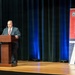 A man in a dark suit and red striped tie is speaking at a wooden podium on a stage. Behind him is a dark teal curtain with blue stage lighting, and to his right stands a banner for the 'Department of War Manufacturing Technology Program' marking its 70th anniversary. The event appears to be a formal ceremony or conference.