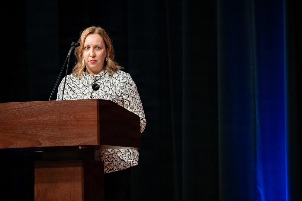 A woman with blonde hair is standing at a wooden podium and speaking into a microphone at what appears to be a formal event or conference. She is wearing a black and white patterned jacket with a dark brooch. The background features dark curtains with blue stage lighting on the right side.