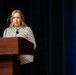 A woman with blonde hair is standing at a wooden podium and speaking into a microphone at what appears to be a formal event or conference. She is wearing a black and white patterned jacket with a dark brooch. The background features dark curtains with blue stage lighting on the right side.