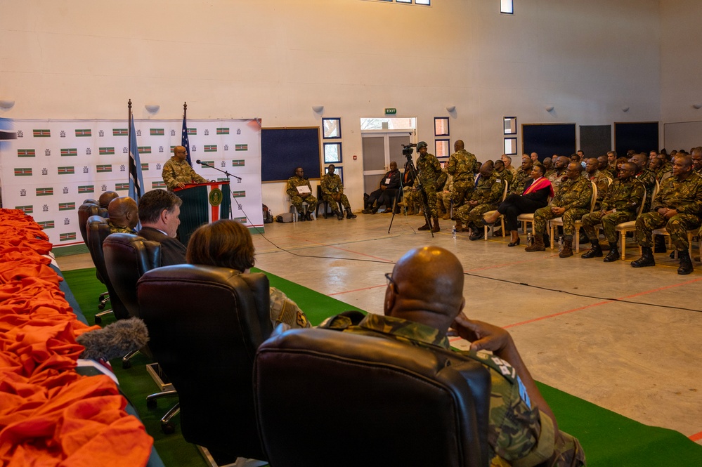 A formal military ceremony or briefing is taking place in a large indoor hall, with a speaker at a podium addressing a seated audience of uniformed military personnel. Two flags are visible behind the podium alongside a branded backdrop featuring what appears to be national insignia. A mix of military and civilian dignitaries are seated in the front row facing the speaker.
