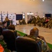 A formal military ceremony or briefing is taking place in a large indoor hall, with a speaker at a podium addressing a seated audience of uniformed military personnel. Two flags are visible behind the podium alongside a branded backdrop featuring what appears to be national insignia. A mix of military and civilian dignitaries are seated in the front row facing the speaker.