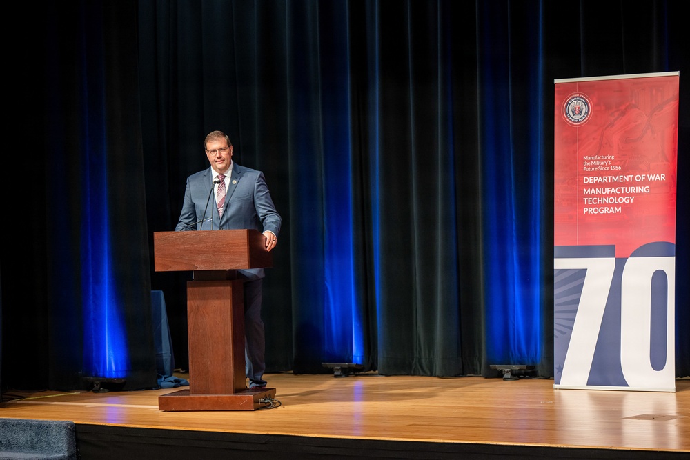 A man in a gray suit is speaking at a wooden podium on a stage. Behind him is a red banner for the 'Department of War Manufacturing Technology Program,' noting it has been 'Manufacturing the Military's Future Since 1956' and displaying the number '70,' suggesting a 70th anniversary event. The stage is set with dark curtains and blue accent lighting.