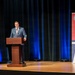 A man in a gray suit is speaking at a wooden podium on a stage. Behind him is a red banner for the 'Department of War Manufacturing Technology Program,' noting it has been 'Manufacturing the Military's Future Since 1956' and displaying the number '70,' suggesting a 70th anniversary event. The stage is set with dark curtains and blue accent lighting.