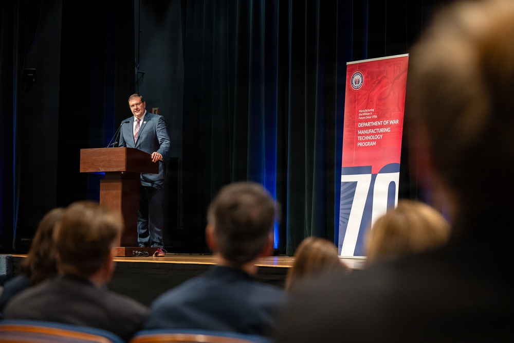 A man in a gray suit is delivering a speech at a wooden podium on a stage before an audience. Behind him stands a red and blue banner for the 'Department of War Manufacturing Technology Program,' featuring the number '70,' suggesting a 70th anniversary event. The setting appears to be a formal auditorium or conference hall.