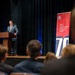 A man in a gray suit is delivering a speech at a wooden podium on a stage before an audience. Behind him stands a red and blue banner for the 'Department of War Manufacturing Technology Program,' featuring the number '70,' suggesting a 70th anniversary event. The setting appears to be a formal auditorium or conference hall.
