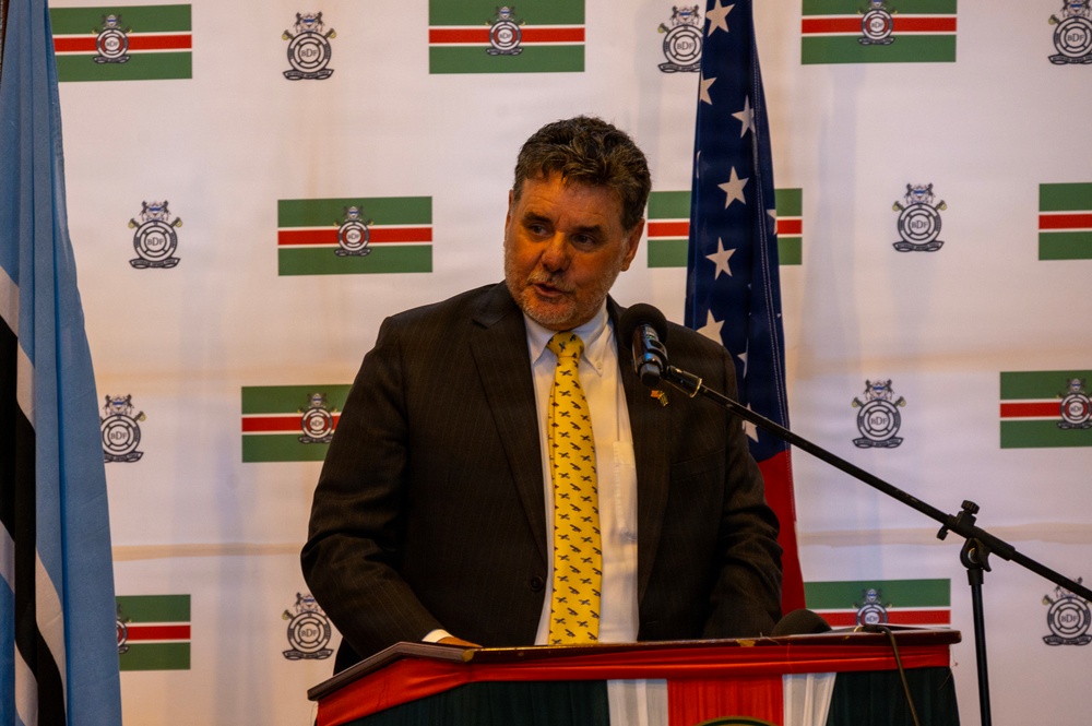 A man in a dark suit and yellow tie is speaking at a red podium with a microphone. Behind him is a branded backdrop featuring a flag with green, white, and red horizontal stripes along with an institutional crest/logo. A United States flag and what appears to be another national or organizational flag are visible to his sides.