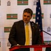 A man in a dark suit and yellow tie is speaking at a red podium with a microphone. Behind him is a branded backdrop featuring a flag with green, white, and red horizontal stripes along with an institutional crest/logo. A United States flag and what appears to be another national or organizational flag are visible to his sides.