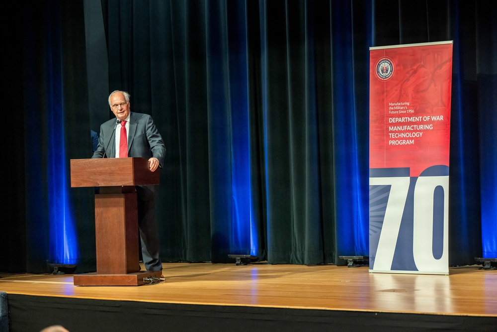 A man in a dark suit and red tie is speaking at a wooden podium on a stage. Behind him is a dark curtain with blue lighting, and to his right stands a red and navy banner for the 'Department of War Manufacturing Technology Program' celebrating its 70th anniversary, with the tagline 'Manufacturing the Military's Future Since 1956'.