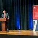 A man in a dark suit and red tie is speaking at a wooden podium on a stage. Behind him is a dark curtain with blue lighting, and to his right stands a red and navy banner for the 'Department of War Manufacturing Technology Program' celebrating its 70th anniversary, with the tagline 'Manufacturing the Military's Future Since 1956'.